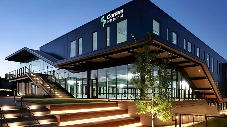 The CordenPharma building at dusk with dark cladding, large glass windows, and illuminated entrance steps